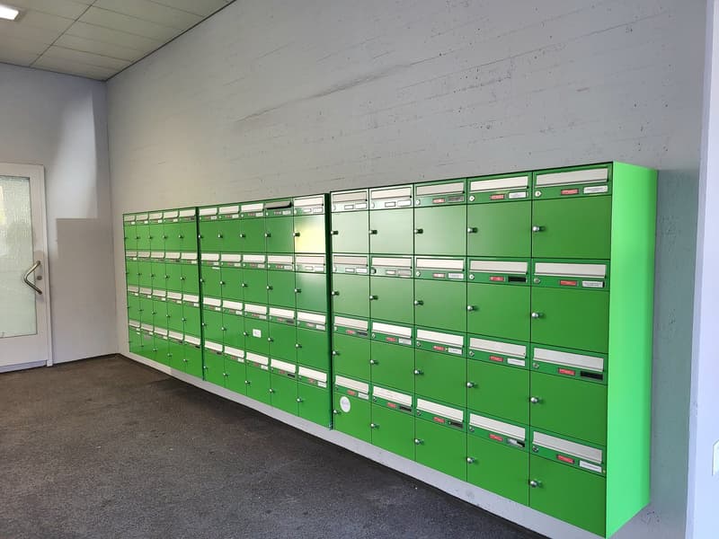 Green mailboxes are lined up on the wall, door, light fixture on ceiling