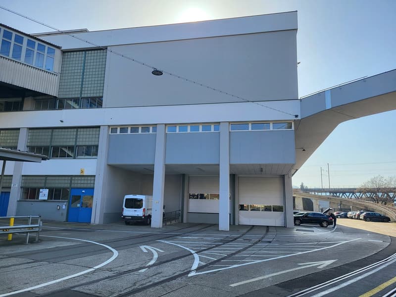 Industrial style building, several windows, blue door, two garages with two vehicles parked, several cars parked in the surrounding area.