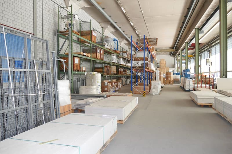 Large storage space with shelves and stacks of materials on wooden pallets. A metal mesh gate and a blue steel gate on the left. Grey cement flooring.