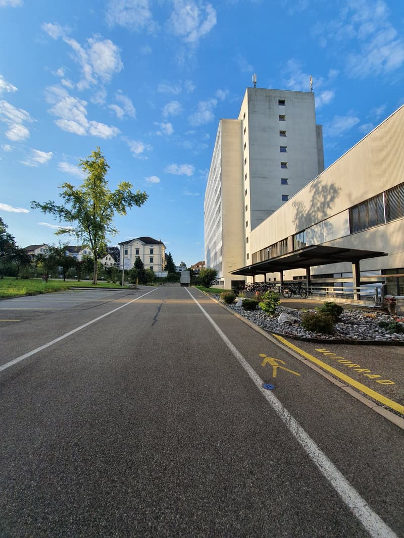 The image shows a large, multi-story concrete building with a modern architectural design. The building is surrounded by a paved area with a bike rack and some landscaping. There are trees and other buildings visible in the background, indicating this is