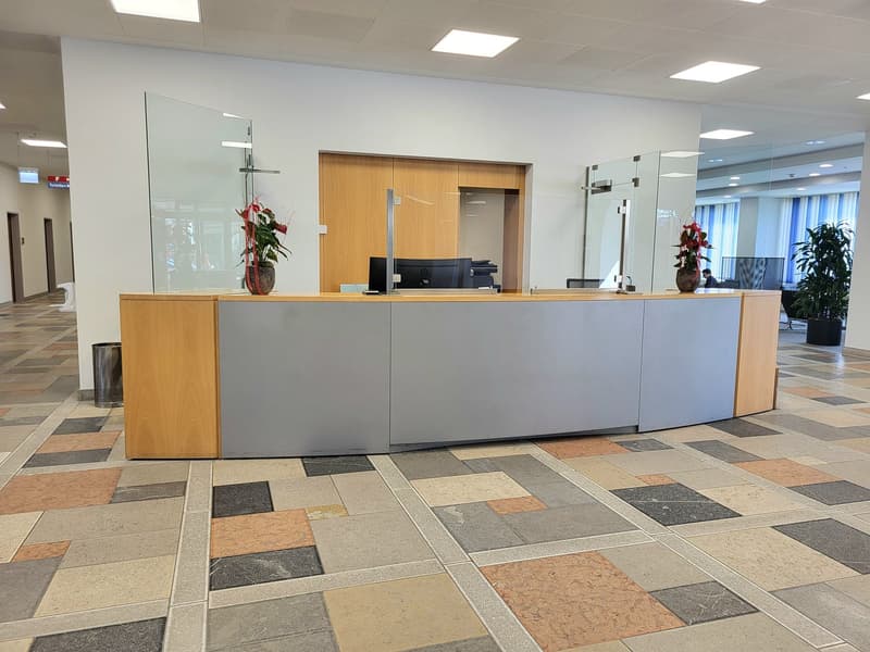 Modern reception desk with glass partitions, wooden front, gray sides, computer setup, flower vases, polished floor, trash can, office environment.