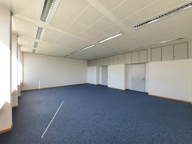 Empty room with blue carpeted floor, white walls, and recessed lighting. The ceiling has a grid design and metal grates. There are cabinets and doors along the wall.