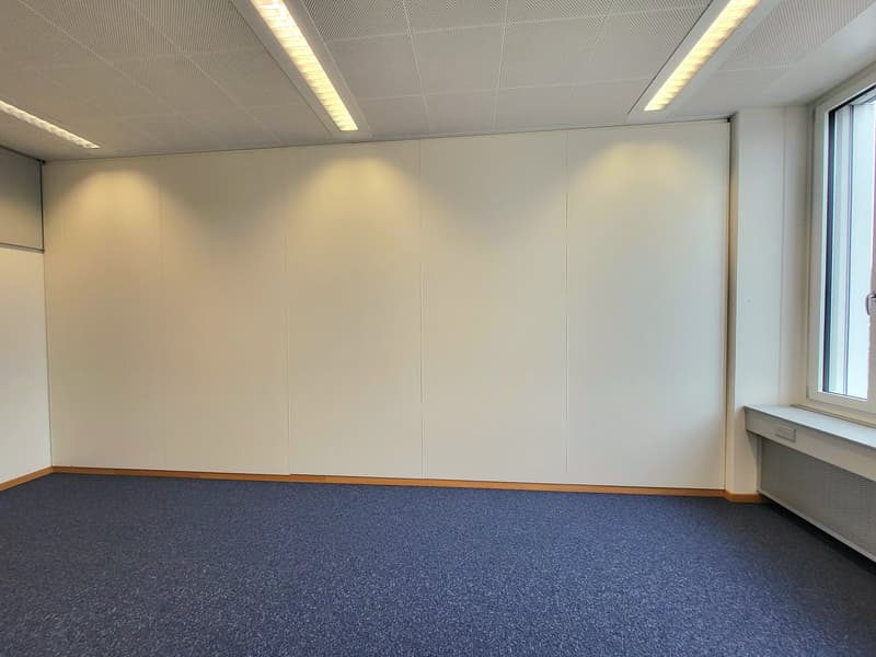 The image shows a spacious, modern office space with white walls, a suspended ceiling with recessed lighting, and a large window. The floor is covered in a blue carpet. The room appears to be empty, ready for use as an office or meeting space.