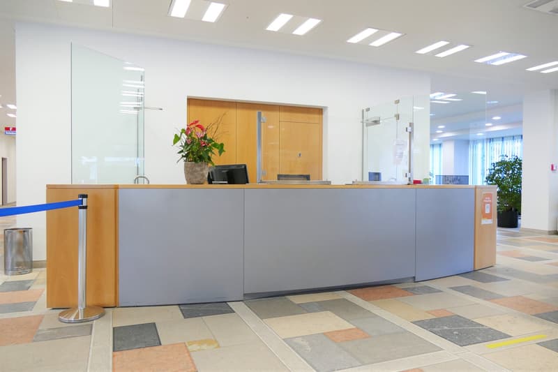Bright and modern office lobby with a large reception desk made of wood and gray panels. The desk has a computer monitor and a vase of flowers. The floor is tiled with a patterned design. The lobby has large windows and bright lighting from the ceiling.