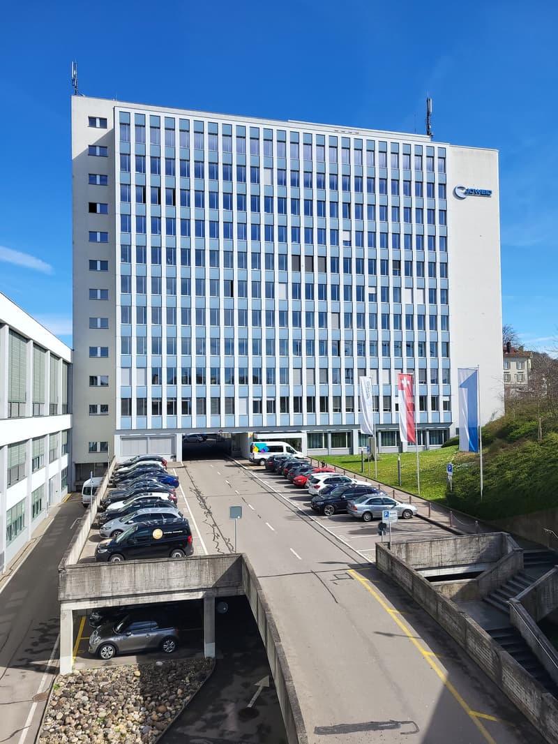 A large multi-story office building with a parking lot in the foreground. The building has a modern, rectangular design with many windows. The parking lot appears to have several cars parked in it.