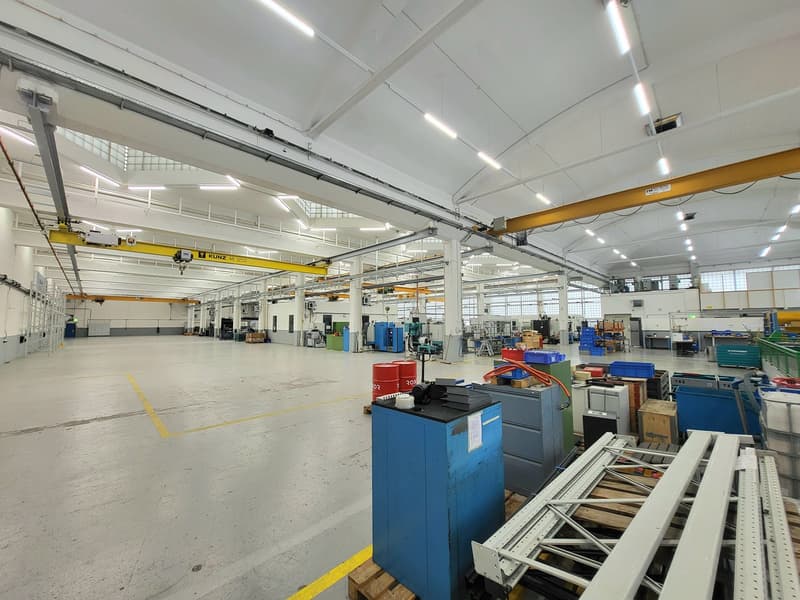 Large industrial workshop with high ceilings, overhead cranes, and various machinery and equipment. The space appears to be used for manufacturing or assembly work, with storage racks and workstations visible throughout the open floor plan.