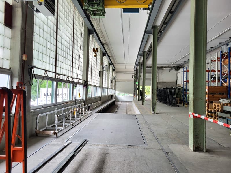 Large industrial warehouse space with concrete floors, metal shelving, and overhead cranes. The space appears to be used for storage and distribution, with various equipment and materials visible.