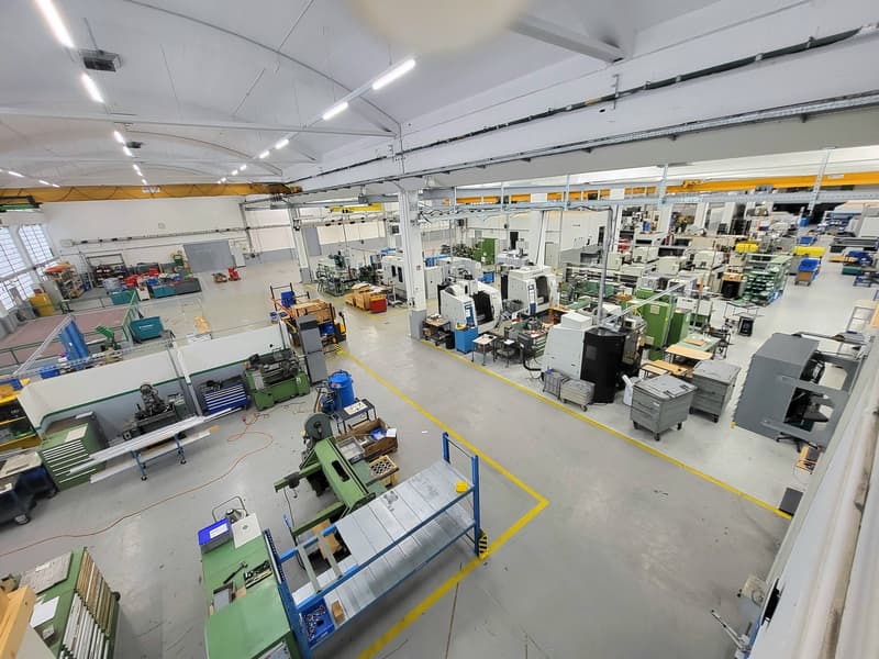The image shows a large industrial workshop or manufacturing facility. The space is filled with various machinery, tools, and equipment used for production or assembly. The workshop has high ceilings, overhead lighting, and a clean, organized layout with
