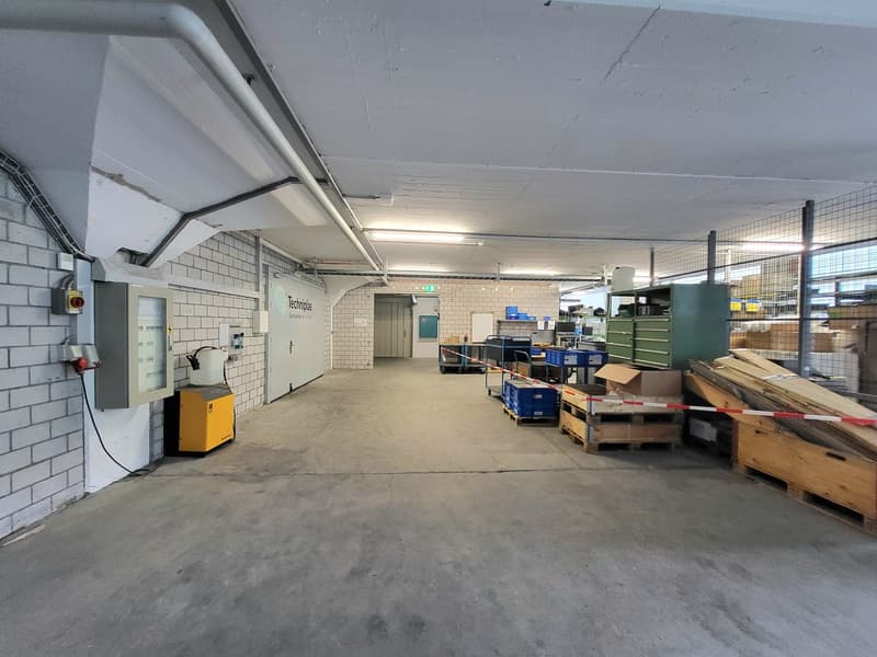 The image shows a large industrial warehouse space with concrete floors, white brick walls, and a high ceiling. There are various storage shelves, boxes, and equipment visible throughout the space, indicating it is used for storage and distribution purpos