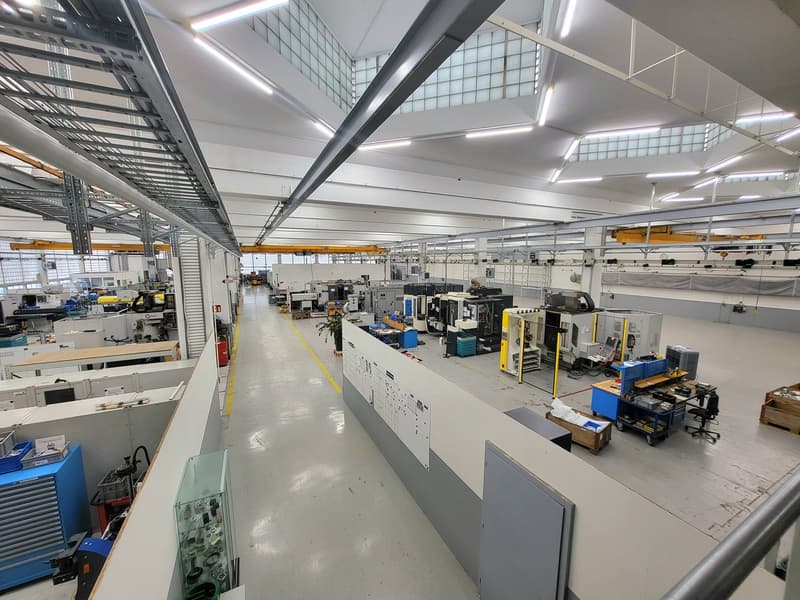 Large industrial workshop with high ceilings, multiple workstations, machinery, and storage racks. The space appears to be used for manufacturing or assembly work, with various tools and equipment visible throughout the facility.