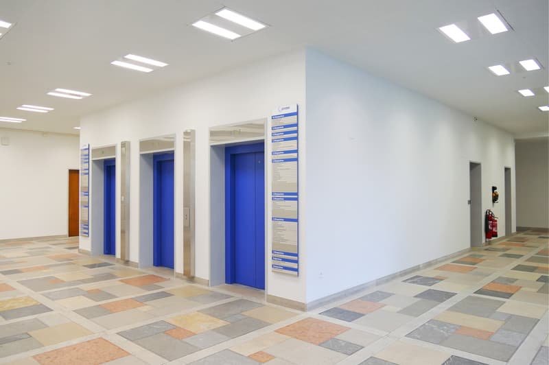 Empty hallway with blue elevator doors and fire extinguishers, tiled floor and white walls
