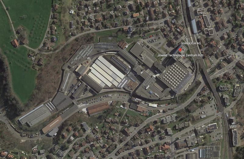 Industrial area with multiple buildings, a factory marked with a red dot, residential houses, green fields, a large open area, roads, parking lots, a railway, and solar panels on some buildings.