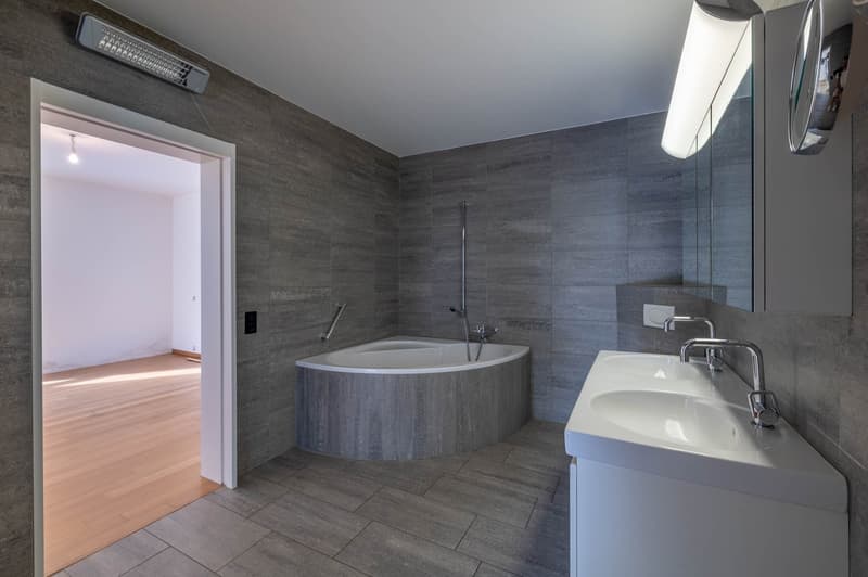 Modern bathroom with grey tiled walls, a freestanding bathtub, a double sink vanity, and built-in storage. The space features sleek lighting and a wooden floor in the adjacent area.