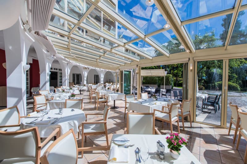modern restaurant with white chairs, glass walls, white tablecloths, white flowers in a vase, tiled floor, large glass windows, glass ceiling