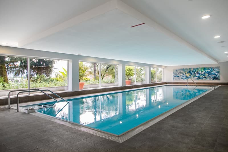 Indoor swimming pool with clear blue water, large windows, handrails, and tiled floor.
