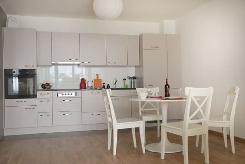 Kitchen with modern white furniture, table, chairs, dishwasher, oven, white backsplash.