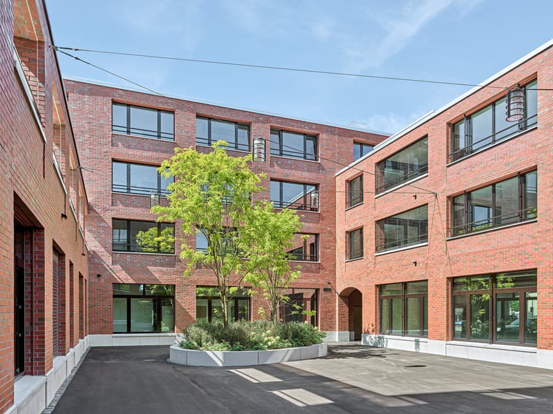 A multi-story brick building with large windows, a central courtyard with a tree, and a paved outdoor area.
