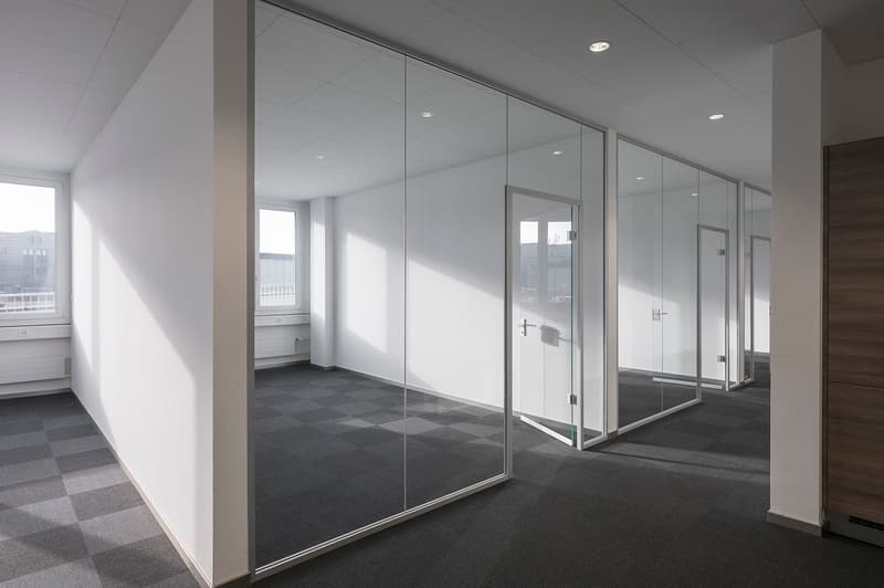 Modern office with white walls, glass partitions, checkered flooring, and windows with outside views.