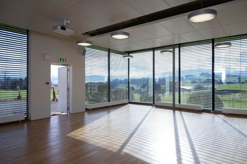 Large open office space with floor-to-ceiling windows providing a scenic view of the outdoor landscape. The room has a hardwood floor, recessed lighting, and a door leading to the exterior. The blinds on the windows are partially open, allowing natural li