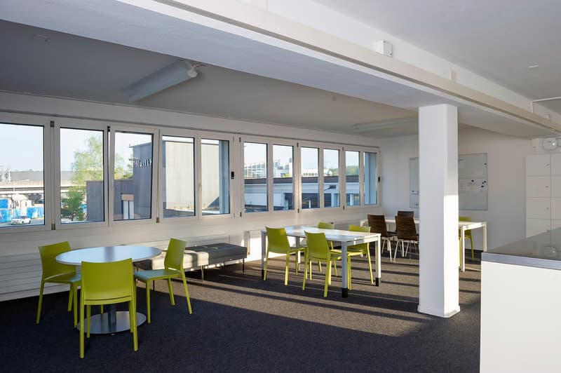 Large open office space with floor-to-ceiling windows, white walls, and gray carpeted flooring. The space features several round tables with green chairs, providing a collaborative work environment.