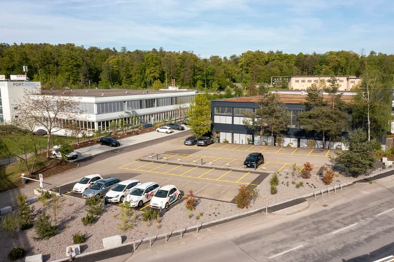 The image shows a commercial property with a large parking lot. The building has a modern, rectangular design with glass windows. There are several cars parked in the lot, which is marked with yellow lines. The surrounding area is wooded, with trees and g