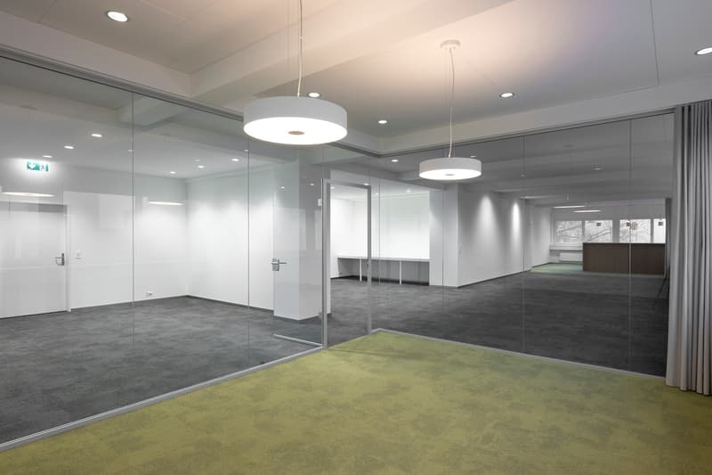 glass walls, green carpeted floor, gray carpeted floor, white walls, white ceiling, open space, ceiling lamps