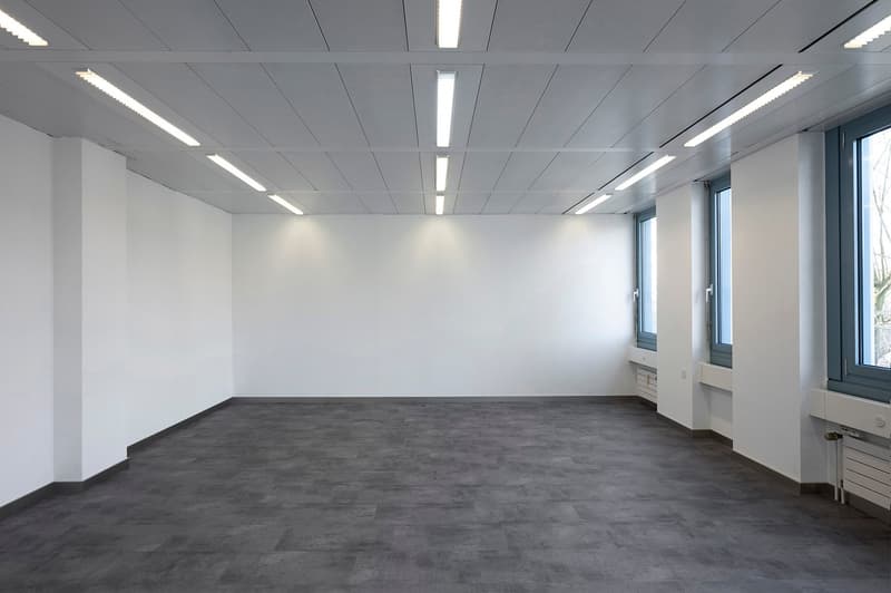 large empty room with white walls, gray floor, and windows, recessed ceiling lights