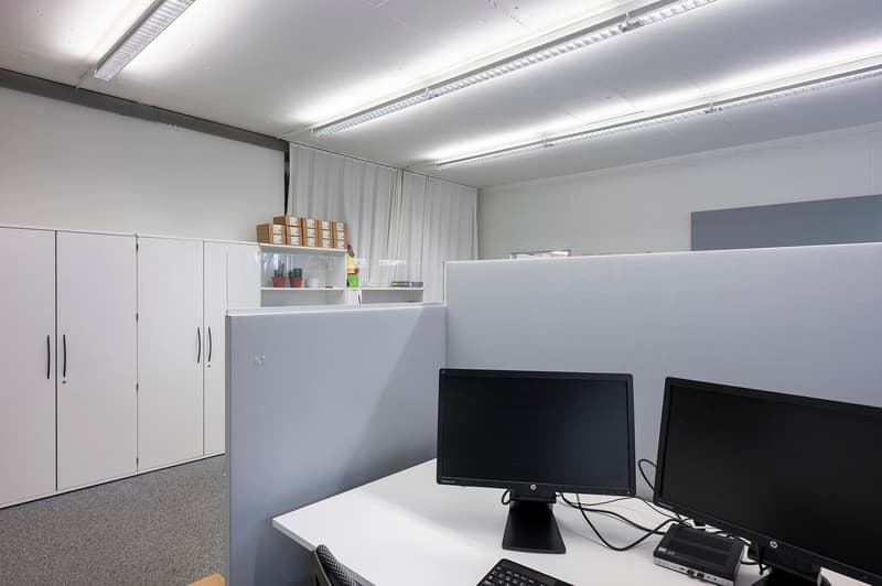 modern office setup with white walls, gray dividers, desk with two monitors, shelves with storage boxes, and a white cupboard