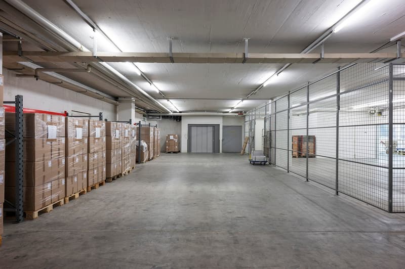 long corridor with boxes, storage shelves, large fenced area, grey walls, grey floor, steel-reinforced doors