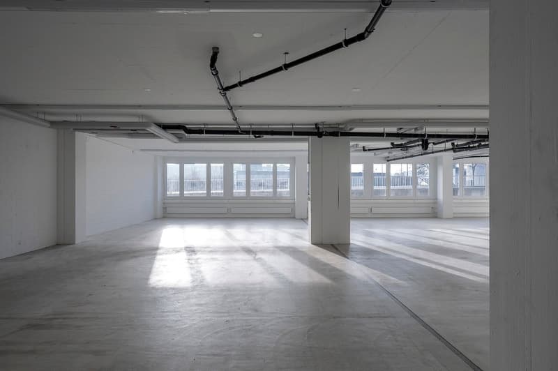An empty room with white walls, gray floor, large windows on the right side, a central pillar, pipes attached to the ceiling and floor