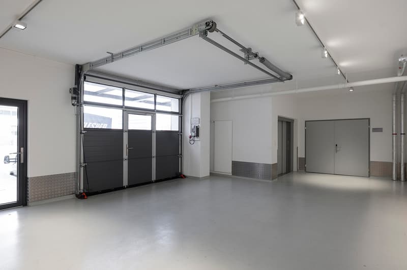 Empty garage with automatic roll-up doors, white walls, gray floor, metal ceiling panels, ceiling mounted lights, door on right side, electrical panel on left side