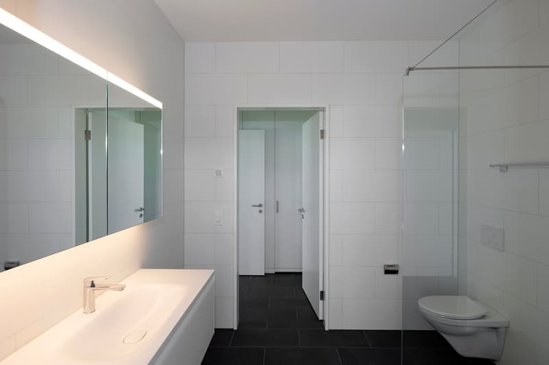 Contemporary bathroom with a sink, large mirror, toilet, and walk-in shower. White tiled walls and floors.