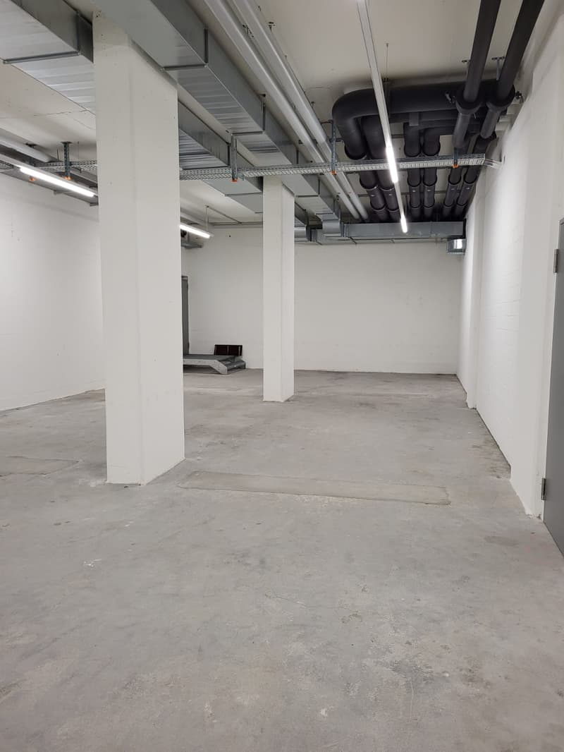 Large open space with concrete flooring, exposed ceiling with ventilation ducts, and white walls. The space appears to be a commercial or industrial area, potentially a warehouse or storage facility.