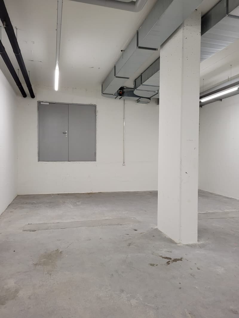 The image shows a large, open space with concrete floors, white walls, and exposed ceiling beams and ductwork. There is a gray metal door visible on the left side of the room. The space appears to be a warehouse or storage facility, with a utilitarian and