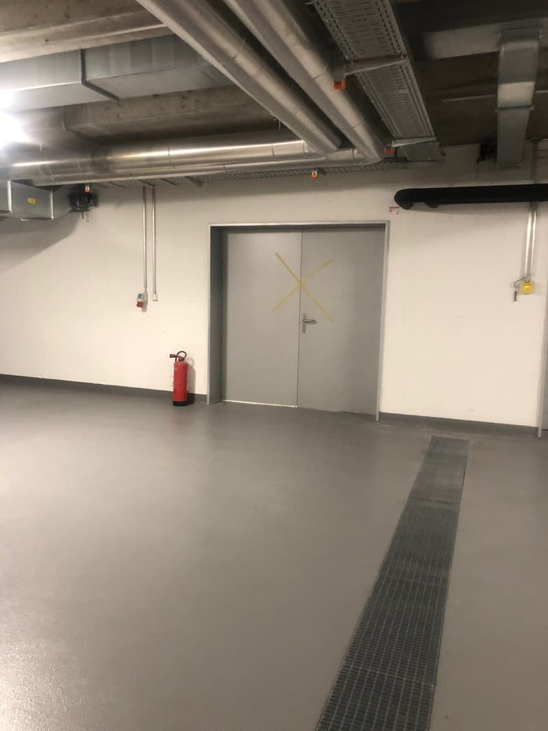 The image shows a large, open garage space with a concrete floor, exposed ceiling beams, and a fire extinguisher mounted on the wall. There is a metal door with an X-shaped design in the center of the frame.