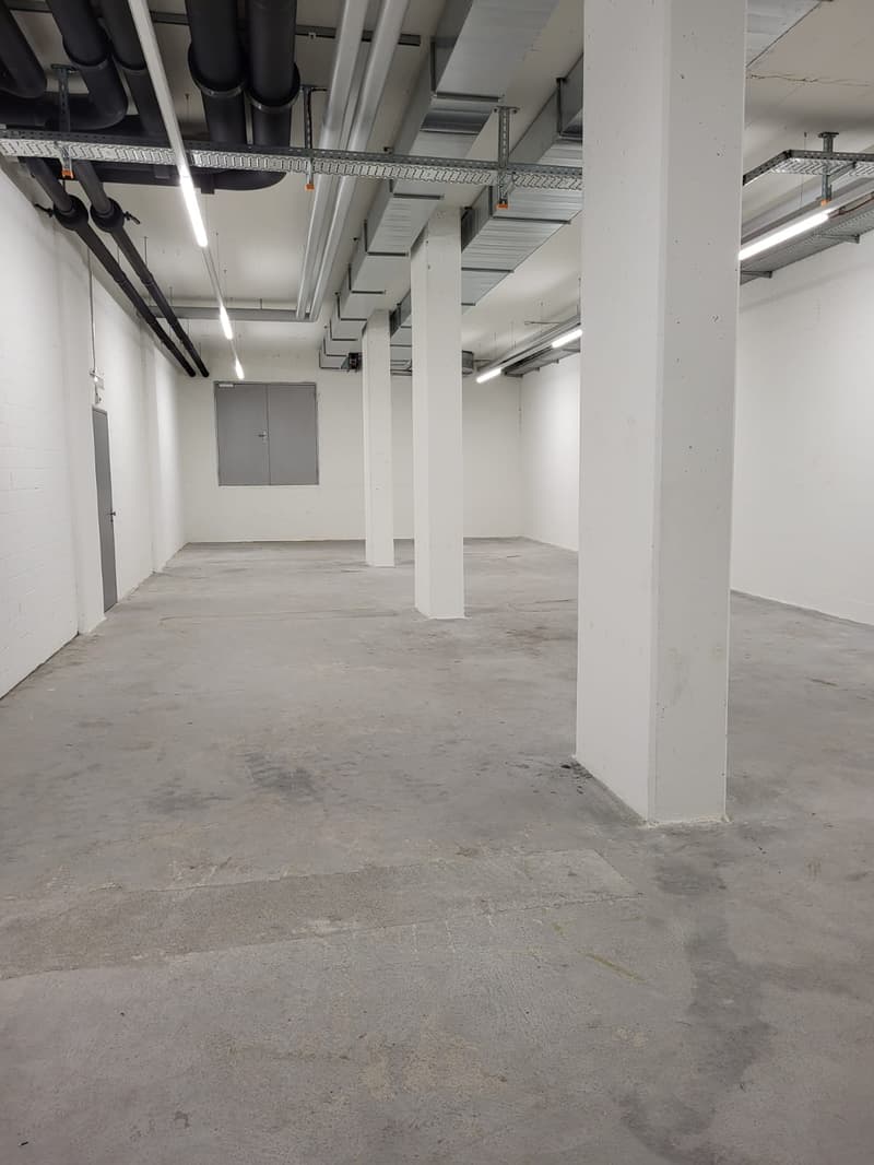 The image shows a large, open warehouse space with concrete floors, white walls, and a high ceiling with exposed ductwork and lighting. The space appears to be empty and unfinished, with a door visible in the background.