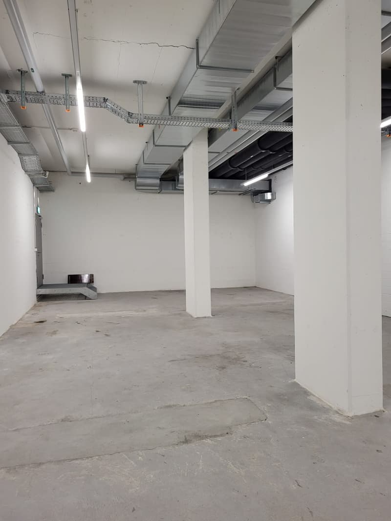 The image shows a large, open commercial space with a concrete floor, white walls, and a high ceiling with exposed ductwork and lighting fixtures. The space appears to be unfinished or under renovation, with no visible furniture or equipment.