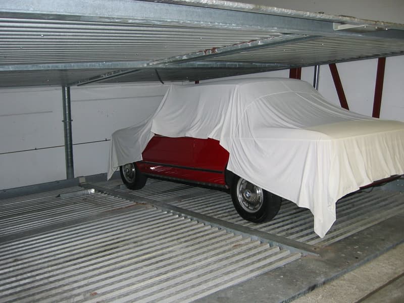 The image shows a covered storage area or garage with a partially covered vehicle inside. The vehicle appears to be a classic or vintage car, with a red body and covered by a white sheet. The storage area has a metal frame and shelving system overhead.