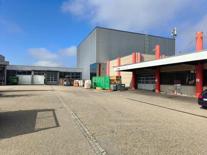 Large industrial building with multiple garages, large open parking area, red pillars, green dumpsters, and a large green container.