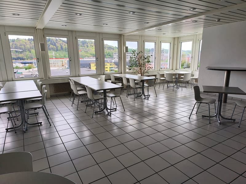 A large room with white tiled floor, white walls, and large windows with a view of the city. There are several tables and chairs arranged in the room.