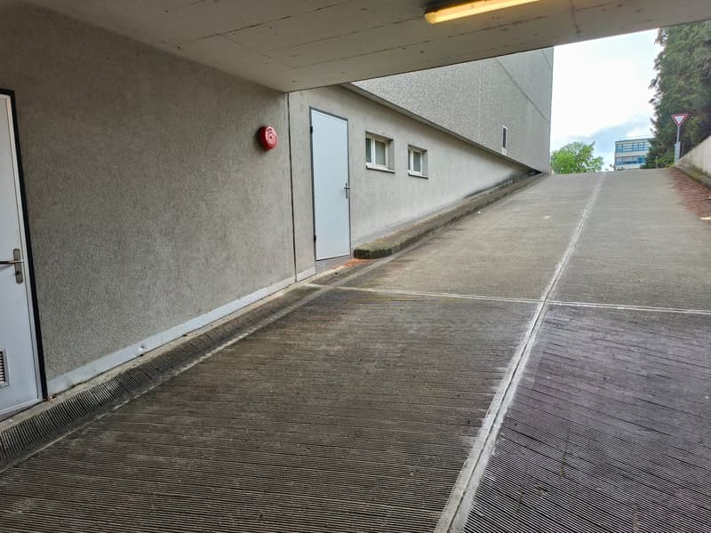 Concrete building with a raised ground floor, white door, red fire extinguisher, and a sloped driveway.
