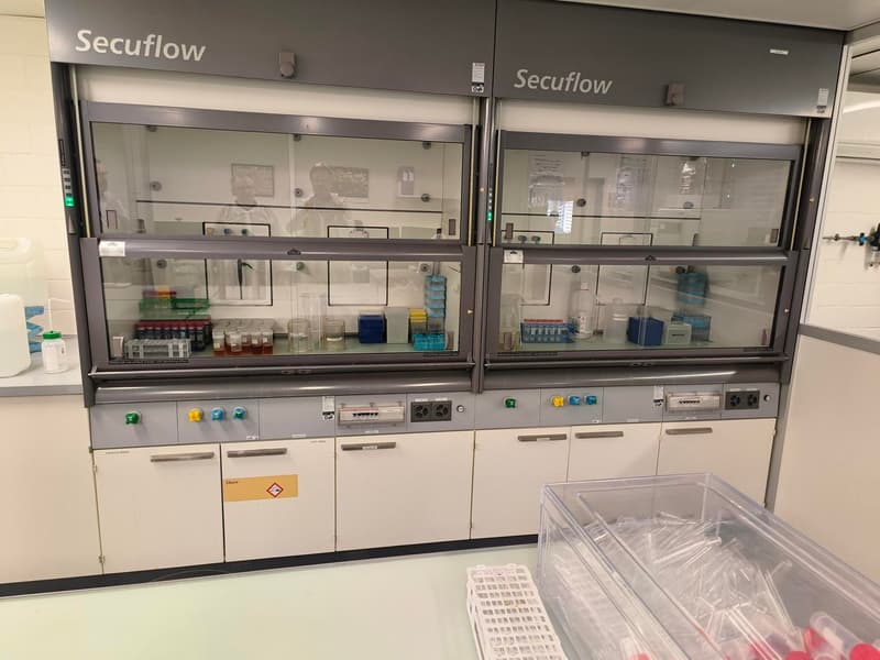Secuflow laboratory equipment with glass enclosures, multiple shelves with lab equipment, white cabinets, and a plastic storage box on the desk.