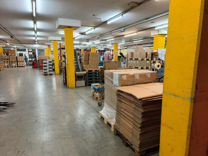 Warehouse with yellow pillars, concrete floor, multiple stacks of boxes, forklifts, and storage equipment.