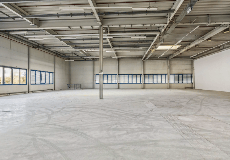 Empty warehouse with white walls, concrete floor, metal beams, and large windows with blue frames.