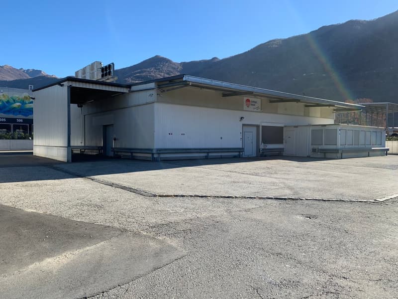 Industrial building with a large parking area in front, white exterior, metal roof, multiple doors, and windows, air conditioning units on the roof, and a sign on the wall.