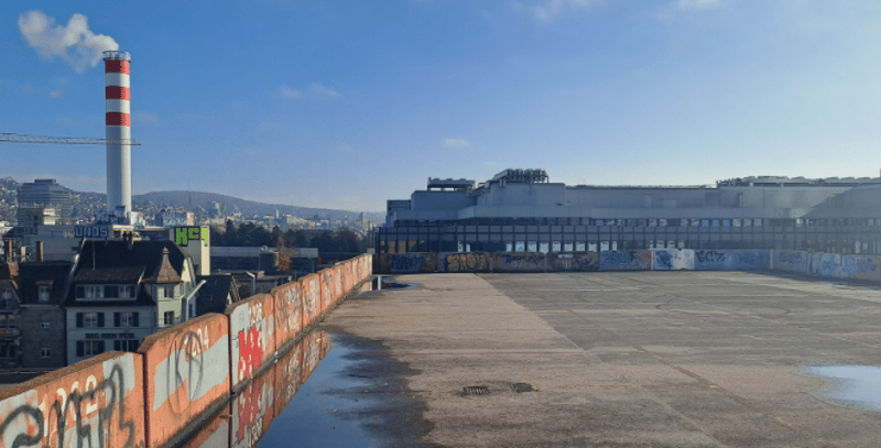 Industrial building with graffiti, large empty lot, industrial chimney, mountains in the background