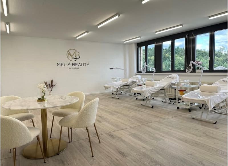 Large, bright room with white walls, wood-look flooring, and several treatment beds and chairs. The room has large windows overlooking a forested area. The Mel's Beauty logo is prominently displayed on the wall.