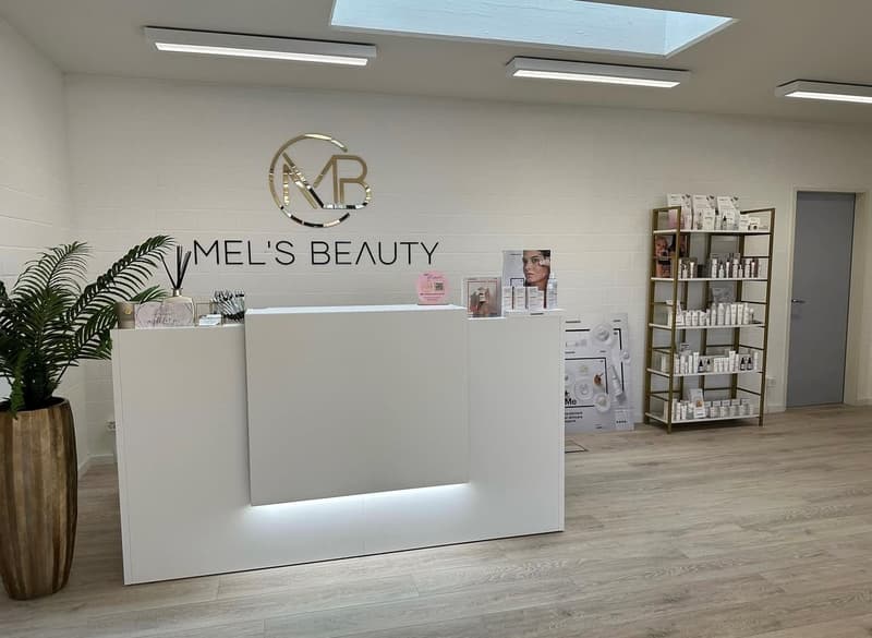 The image shows the interior of a beauty salon called 'Mel's Beauty'. The reception area has a large white counter with the salon's logo displayed prominently. Behind the counter, there are shelves displaying various beauty products. The salon has a moder