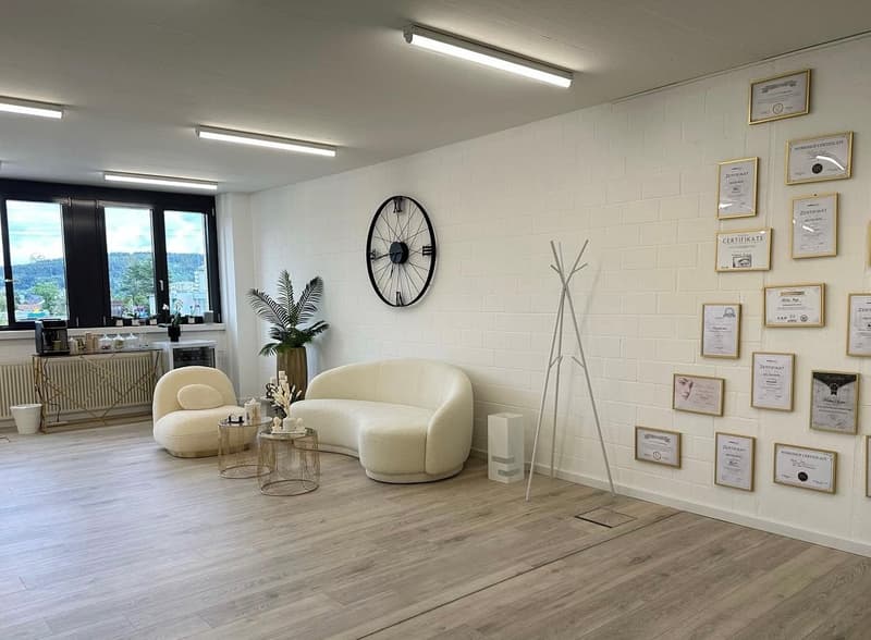 This image shows a modern, well-lit office or studio space with large windows providing a view of the surrounding area. The room has a clean, minimalist design with white walls, hardwood floors, and contemporary furniture including a curved beige sofa and