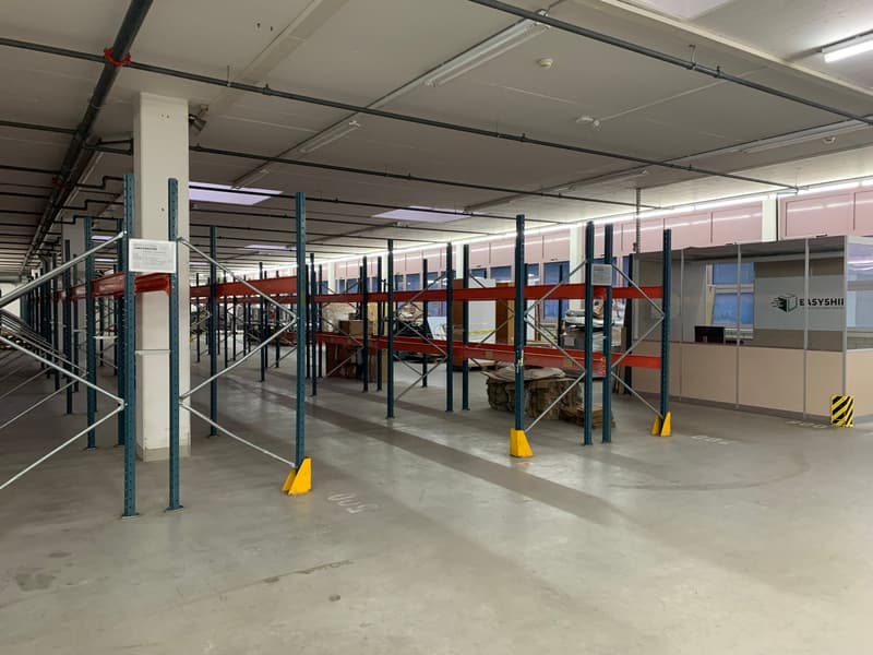 Wide warehouse space with rows of pallet racks, white walls, concrete floor, industrial lighting, various stacked materials and some signage on the wall.